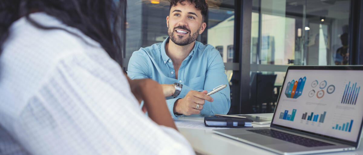 Ein junger Technologieberater sitzt zusammen mit der einer Klientin im Meetingroom und präsentiert Grafiken © courtneyk / Getty Images