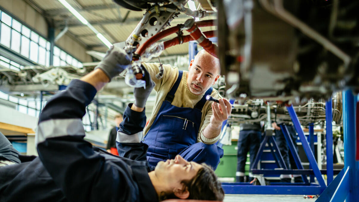 Zwei männliche Flugzeugmechaniker arbeiten an einem Düsentriebwerk in einem Flugzeughangar. Einer von ihnen liegt unter dem Triebwerk, während der andere es mit © Hinterhaus Productions/ Getty Images