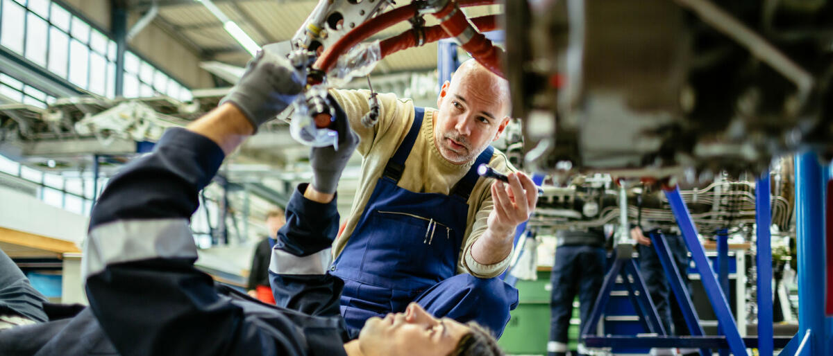 Zwei männliche Flugzeugmechaniker arbeiten an einem Düsentriebwerk in einem Flugzeughangar. Einer von ihnen liegt unter dem Triebwerk, während der andere es mit © Hinterhaus Productions/ Getty Images