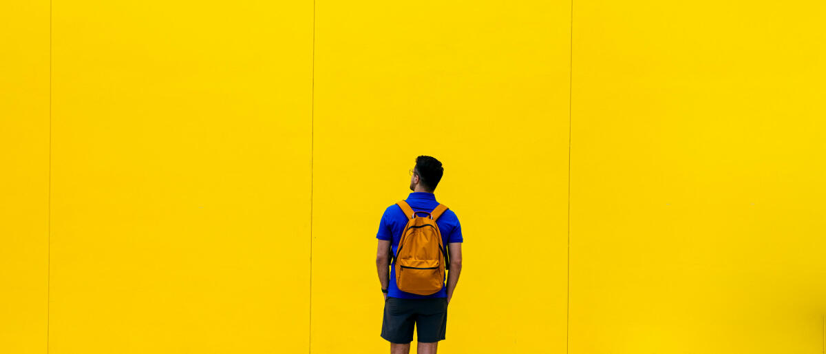 Rückansicht eines Mannes mit Rucksack vor gelber Wand © Alexander Spatari / Getty Images