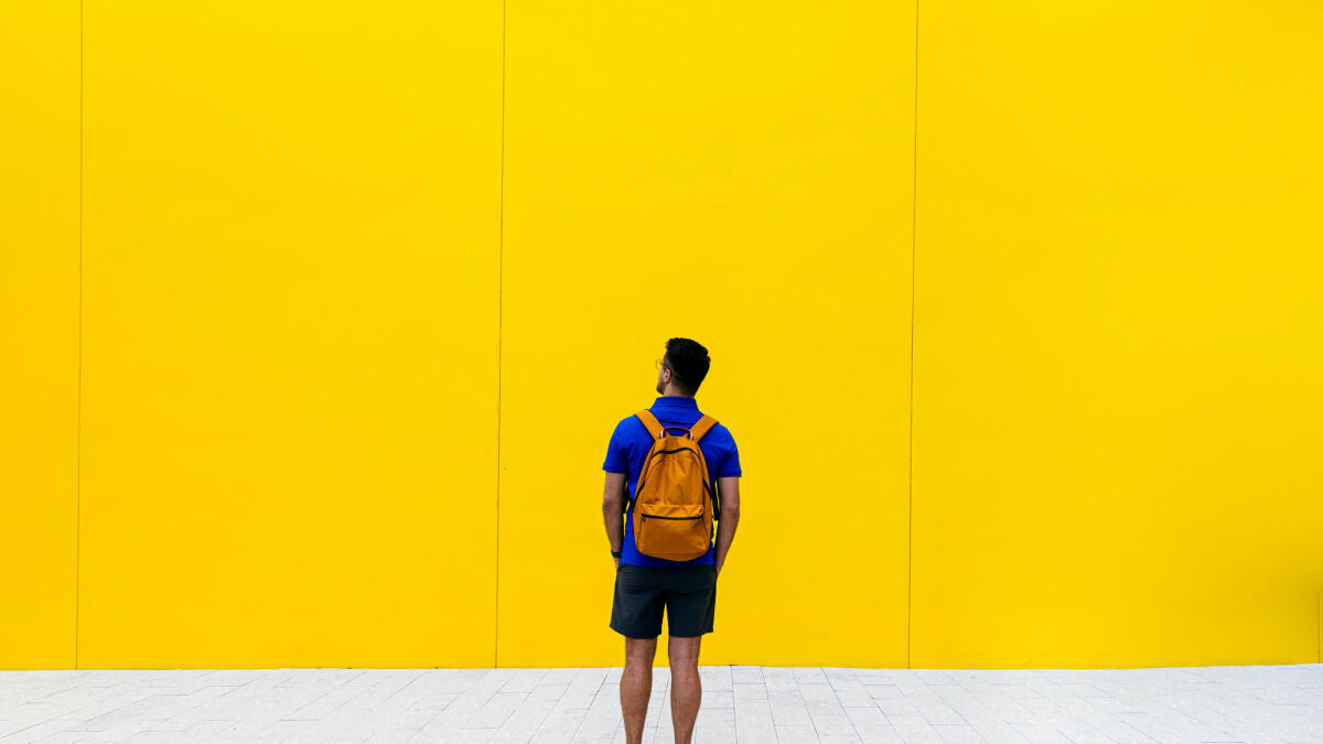 Rückansicht eines Mannes mit Rucksack vor gelber Wand © Alexander Spatari / Getty Images