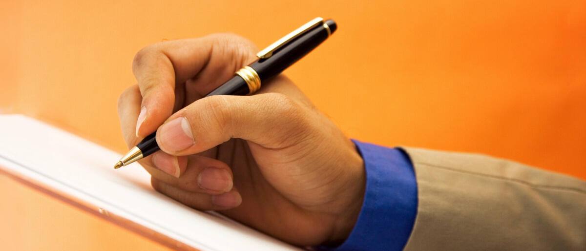 Eine Hand hält einen Kugelschreiber über ein Blatt Papier und setzt zum schreiben an. Der Hintergrund ist einfarbig Orange.  © Nico_Blue / Getty Images