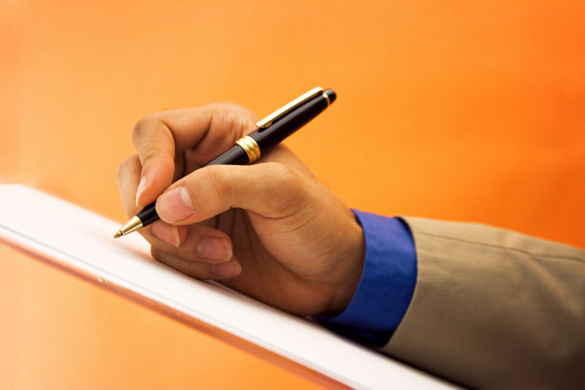 Eine Hand hält einen Kugelschreiber über ein Blatt Papier und setzt zum schreiben an. Der Hintergrund ist einfarbig Orange.  © Nico_Blue / Getty Images