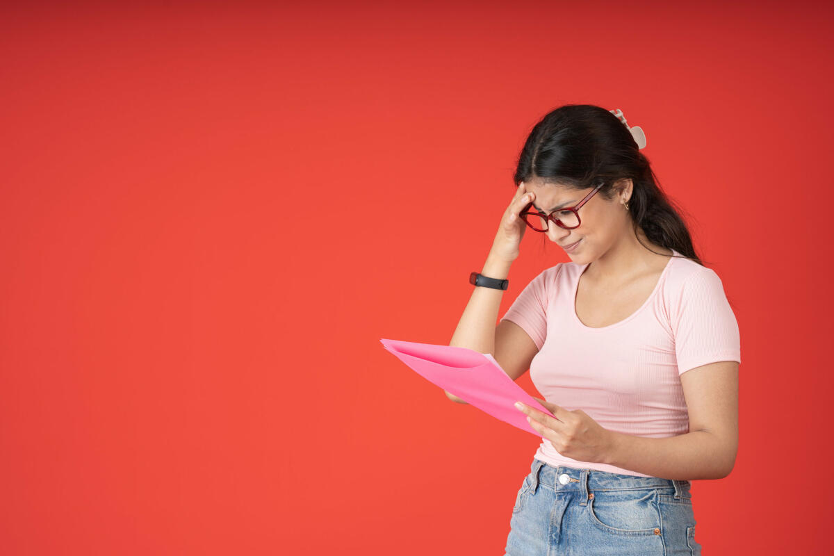 Frau mit Brille, die sich besorgt an den Kopf fasst und einen rosa Ordner mit einer nicht bestandenen Test in der Hand hält, auf rotem Hintergrund © Guillermo Spelucin / Getty Images