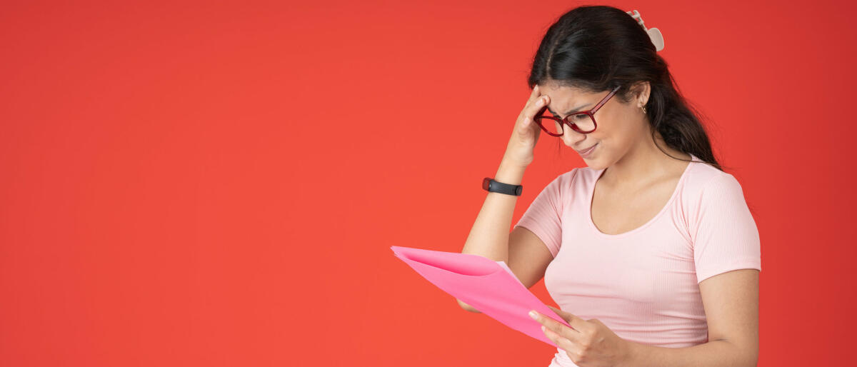 Frau mit Brille, die sich besorgt an den Kopf fasst und einen rosa Ordner mit einer nicht bestandenen Test in der Hand hält, auf rotem Hintergrund © Guillermo Spelucin / Getty Images