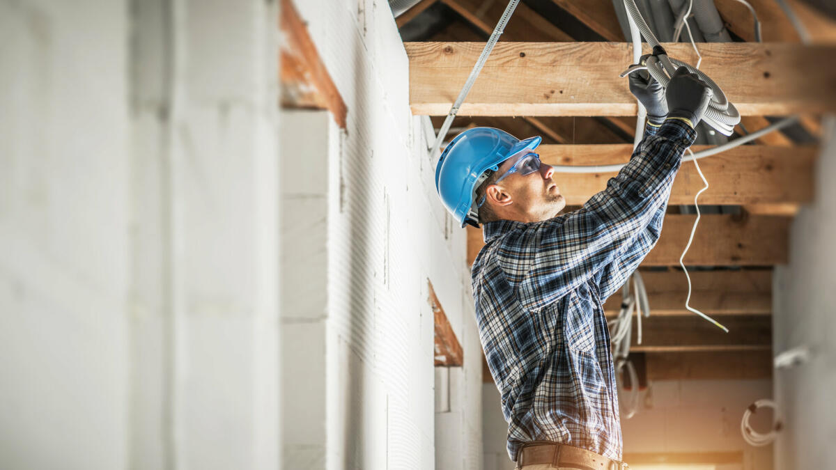 Professioneller kaukasischer Elektriker, der an der Installation der elektrischen Leitungen in der Decke eines neuen Wohnhauses arbeitet. © welcomia/ Getty Images