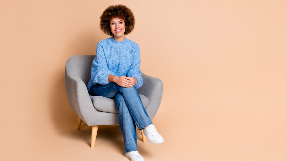 Foto von positivem Mädchen gekleidet strick Pullover Jeans sitzen auf Sessel in der Nähe von Rabatt leerer Raum isoliert auf pastellfarbenem Hintergrund © Deagreez / Getty Images