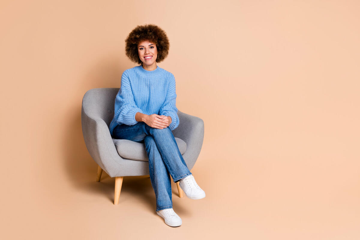 Foto von positivem Mädchen gekleidet strick Pullover Jeans sitzen auf Sessel in der Nähe von Rabatt leerer Raum isoliert auf pastellfarbenem Hintergrund © Deagreez / Getty Images