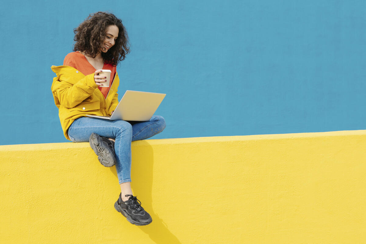 Glückliche junge Frau mit Kaffee zum Mitnehmen, die an einer gelben Wand sitzt und einen Laptop benutzt © Westend61 / Getty Images