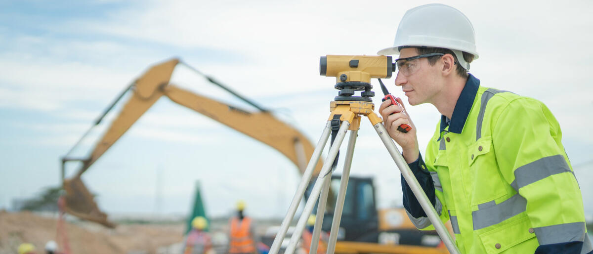 Männlicher Ingenieur bei der Arbeit mit Vermessungsgeräten auf der Baustelle und Arbeiter im Hintergrund © Vithun Khamsong / Getty Images