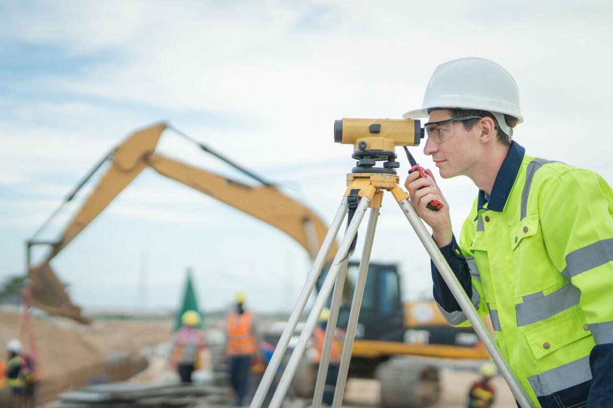 Männlicher Ingenieur bei der Arbeit mit Vermessungsgeräten auf der Baustelle und Arbeiter im Hintergrund © Vithun Khamsong / Getty Images