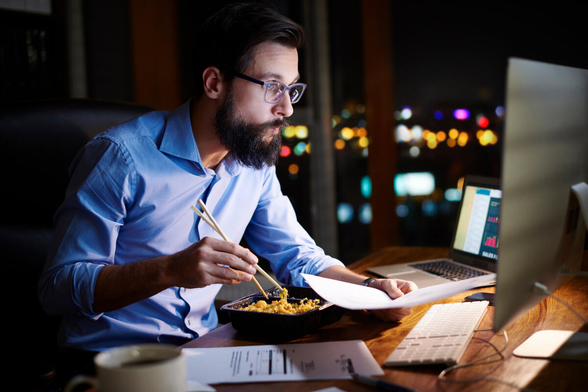 Ein Mann isst nachts vor seinem Computer und macht Überstunden © Gpointstudio / Getty Images