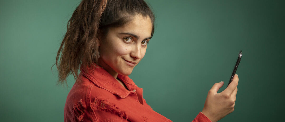 Junge Frau mit Smartphone in der Hand, die kokett in die Kamera guckt und vor einem dunkelgrünen Hintergrund steht. © Westend61 / Getty Images