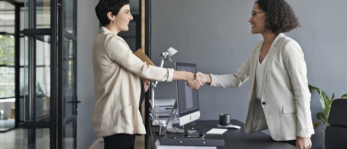 Zwei Frauen geben sich im Büro über den Schreibtisch hinweg die Hand. © Klaus Vedfelt / Getty Images