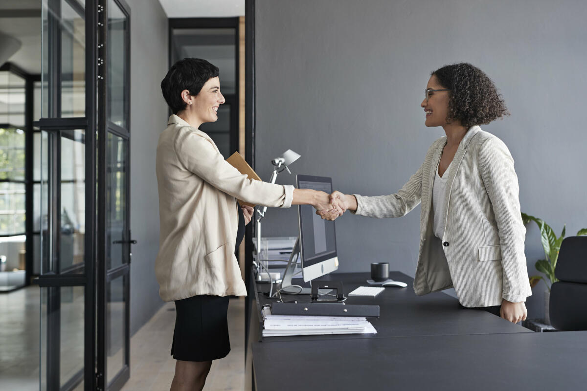 Zwei Frauen geben sich im Büro über den Schreibtisch hinweg die Hand. © Klaus Vedfelt / Getty Images