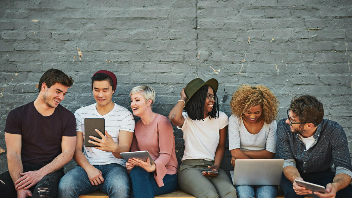 Junge Menschen sitzen gemeinsam auf einer Bank und gucken auf Laptops und Tablets. © PeopleImages / Getty Images