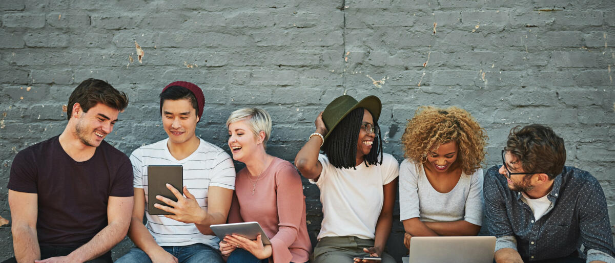 Junge Menschen sitzen gemeinsam auf einer Bank und gucken auf Laptops und Tablets. © PeopleImages / Getty Images