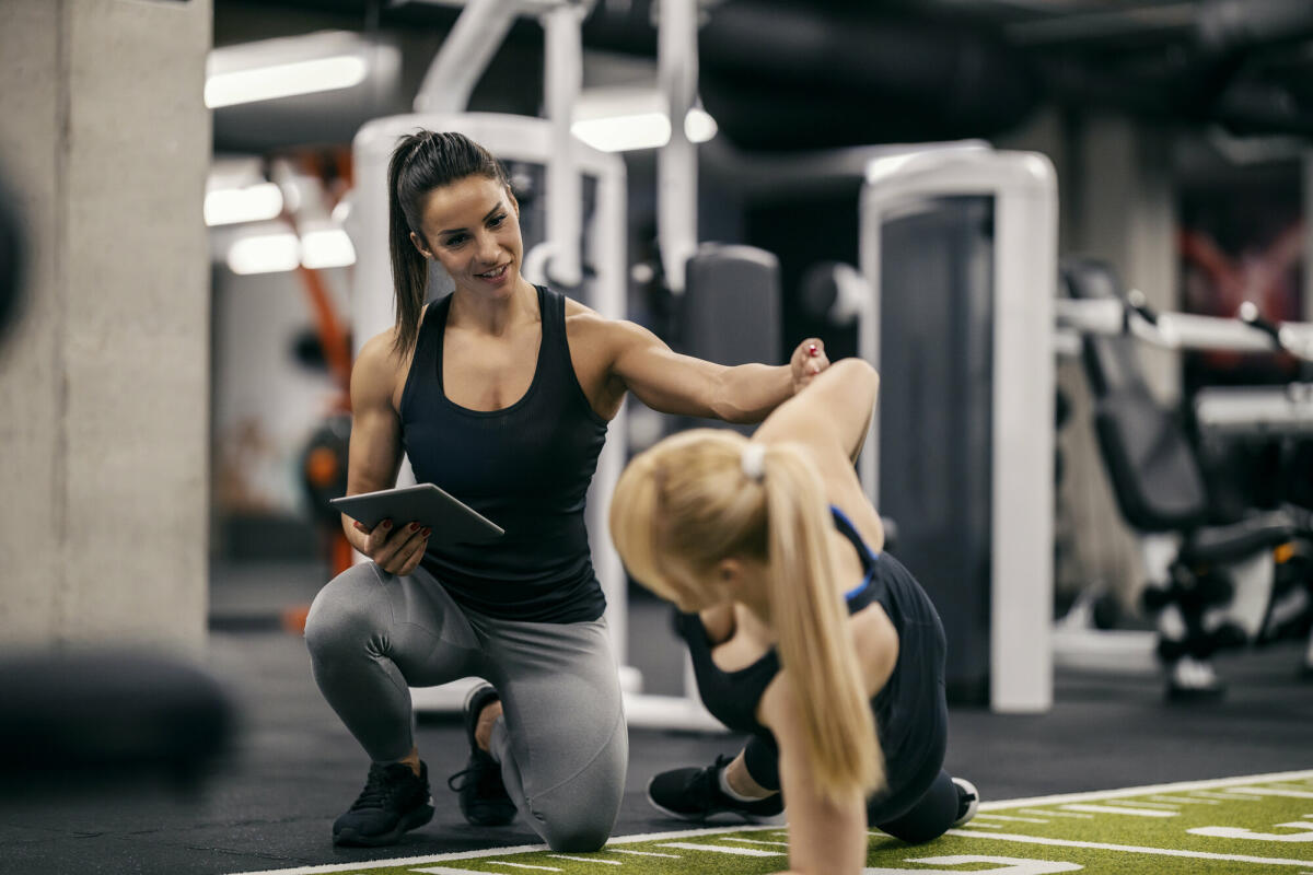 Eine Personal Trainerin trainiert eine Sportlerin in einem Fitnessstudio und verfolgt ihre Fortschritte auf einem Tablet. © dusanpetkovic/ Getty Images